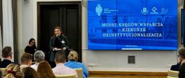 Wiceminister Piechna-Więckiewicz stoi za mównicą i mówi do mikrofonu, obok niej na ścianie wbudowany w ścianę wielki ekranu z napisem Model Kręgów Wsparcia - Kierunek Deinstytucjonalizacji, przed nią sala wypełniona ludźmi.