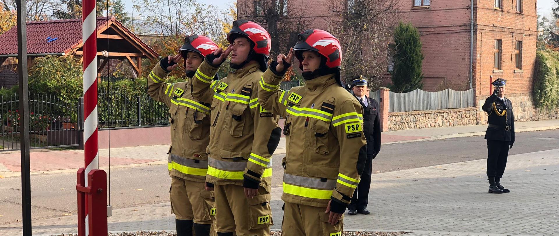 Poczet flagowy złożony ze strażaków PSP podnosi flagę państwową na maszt. Strażacy salutują.