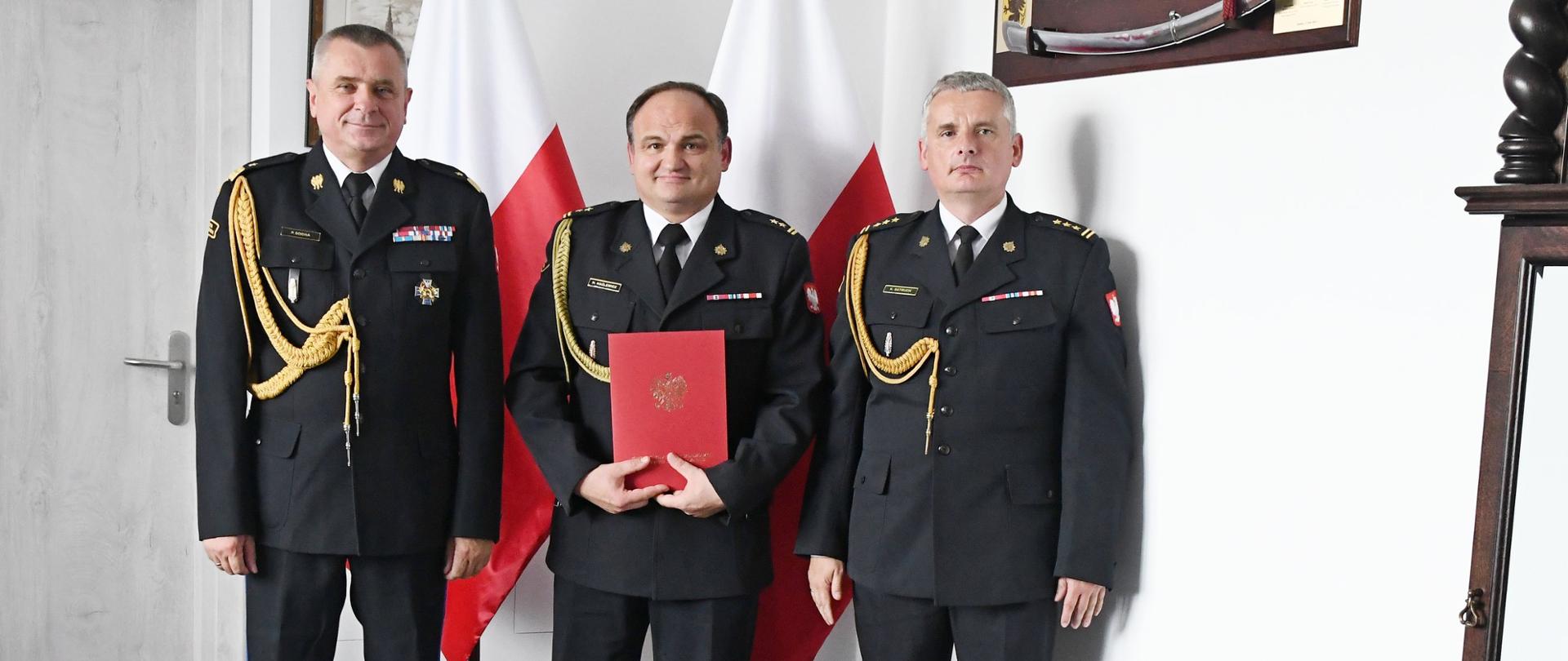 Trzech funkcjonariuszy Państwowej Straży Pożarnej stoi obok siebie strażak w środku trzyma czerwoną teczkę za nimi stoją dwie flagi Polski na ścianie wiszą obraz oraz szabla niedaleko stoi zegar.