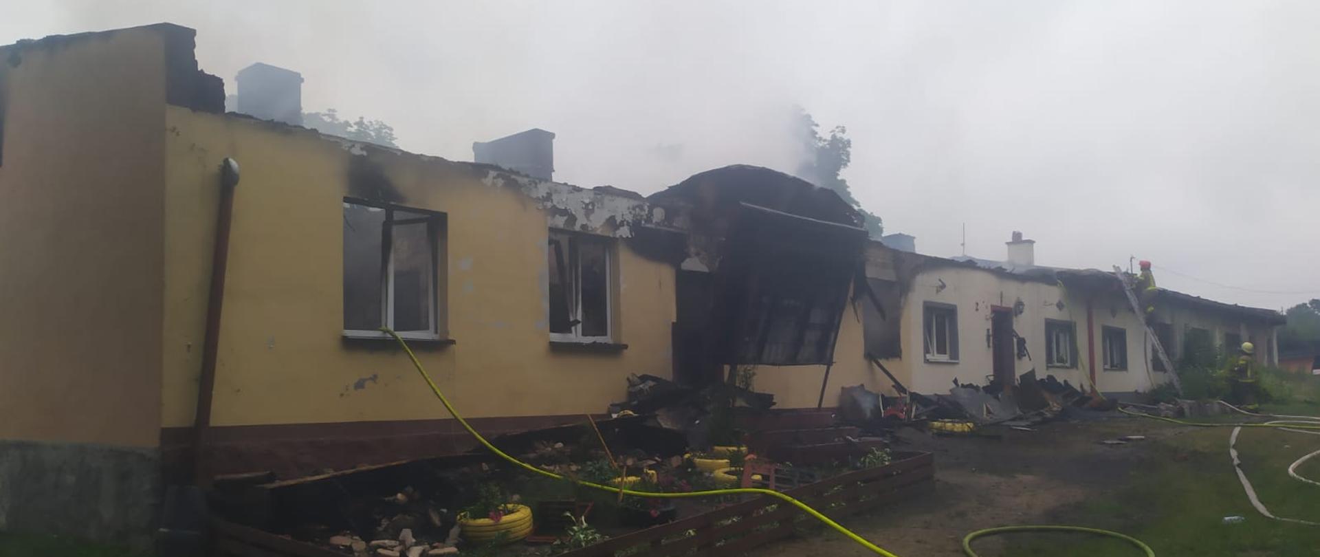 Zdjęcie przedstawia budynek parterowy po pożarze bez dachu. Przed budynkiem rozwinięte są linie gaśnicze.