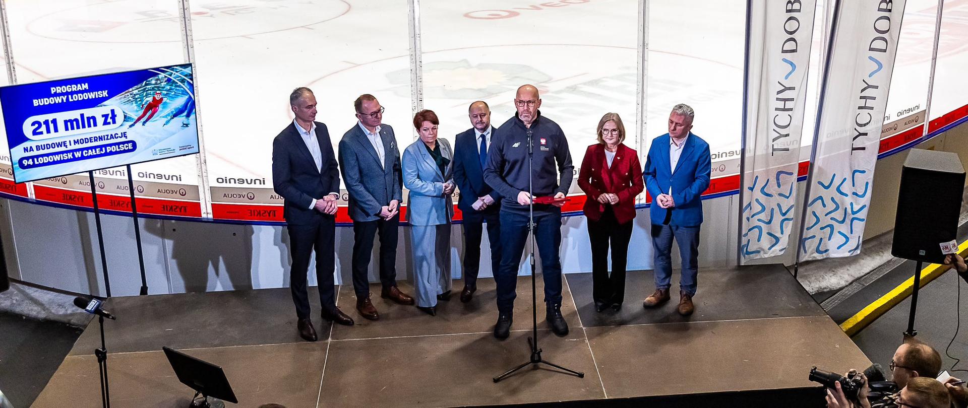 Konferencja prasowa ministra sportu i turystyki Jakuba Rutnickiego. Minister mówi do mikrofonu. Za nim stoją pozostali uczestnicy spotkania. Za nimi widać kryte lodowisko. W prawym dolnym rogu widać fotoreporterów.