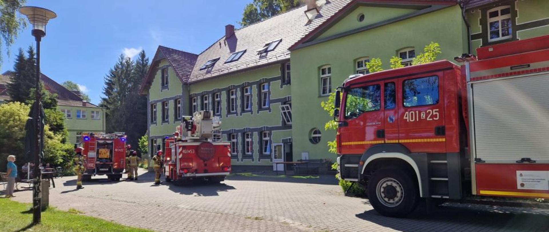 Na placu przed budynkiem stoją trzy wozy strażackie, w tym pojazd z drabiną mechaniczną. Strażacy prowadzą działania przy Domu Pomocy Społecznej, a mieszkańcy obserwują przebieg akcji z bezpiecznej odległości. Słoneczna pogoda podkreśla wyraźne barwy czerwonych pojazdów na tle zielonego budynku. Zdjęcie obrazuje koordynację i skalę ćwiczeń.