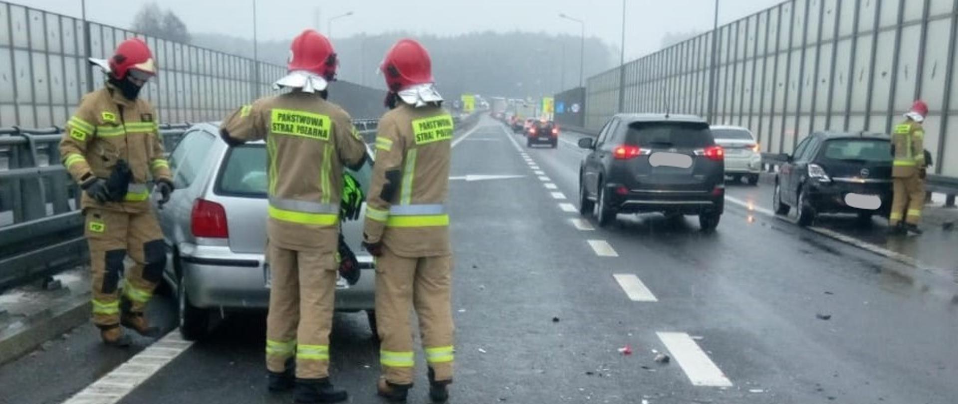 Na zdjęciu widzimy perspektywę drogi ekspresowej S74, na poboczach z lewej i prawej strony znajdują się uszkodzone pojazdy, strażacy prowadzą działania ratownicze.