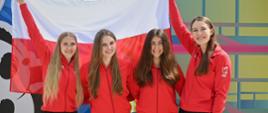 Cztery kobiety w czerwonych sportowych bluzach stoją w szeregu i trzymają za sobą polską flagę.