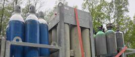 Kilkanaście dużych stalowych butli gazowych poustawianych pionowo na platformie transportowej lub przyczepie. Butle zabezpieczone czerwonym pasem transportowym przymocowanymi do konstrukcji pojazdu. W tle widoczne są drzewa oraz jasne niebo. 