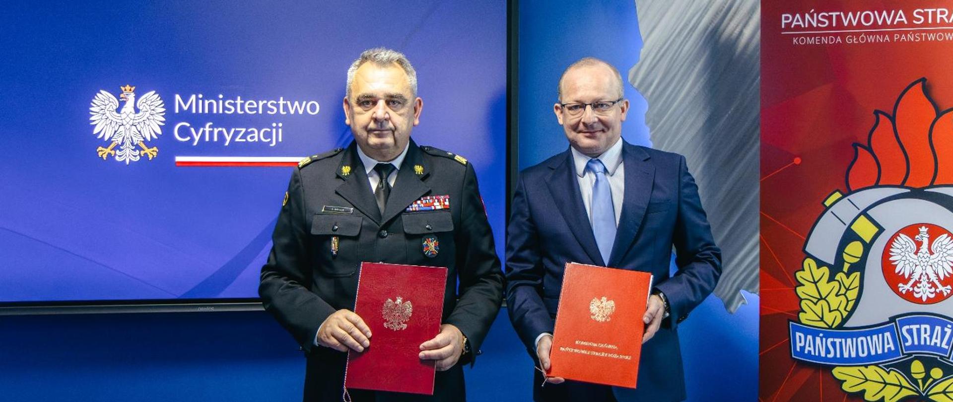 Zastępca Komendanta Głównego Józef Galica i wiceminister cyfryzacji Paweł Olszewski trzymają w ręku czerwone teczki. W tle ekran z napisem Ministerstwo Cyfryzacji oraz baner Państwowej Straży Pożarnej 