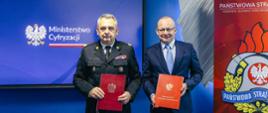 Zastępca Komendanta Głównego Józef Galica i wiceminister cyfryzacji Paweł Olszewski trzymają w ręku czerwone teczki. W tle ekran z napisem Ministerstwo Cyfryzacji oraz baner Państwowej Straży Pożarnej 