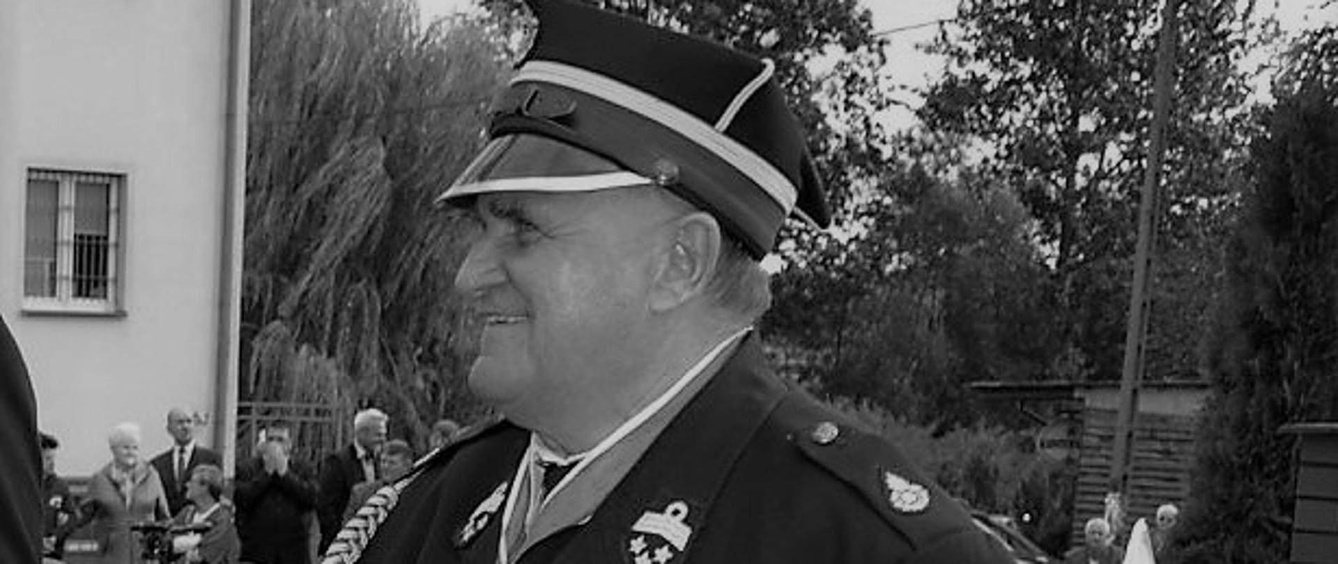 Czarno białe zdjęcie przedstawia strażaka w mundurze galowym ze sznurem i wieloma odznaczeniami – jeden medal zawieszony na szyi, oraz rogatywką na głowie. W tle ludzie zgromadzeni na uroczystości.