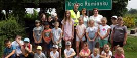 Minister Przemysław Czarnek i dwie kobiety stoją w otoczeniu grupy dzieci. Część z nich stoi, a część siedzi na trawie. Za plecami ministra zielona tablica przypominająca znak drogowy z napisem Kraina Rumianku. Minister Przemysław Czarnek ma na sobie kamizelkę odblaskową. W tle zadaszona altana obrośnięta bujną roślinnością. 