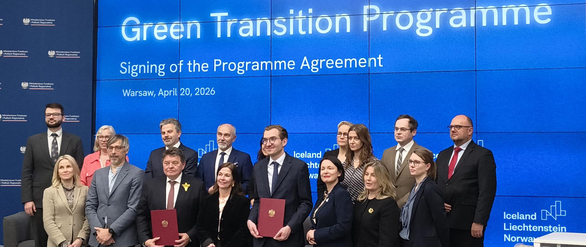 W dużej sali konferencyjnej, na tle niebieskiego ekranu, stoi grupa elegancko ubranych kobiet i mężczyzn. Dwóch mężczyzn trzyma teczki z orłem na okładce. 