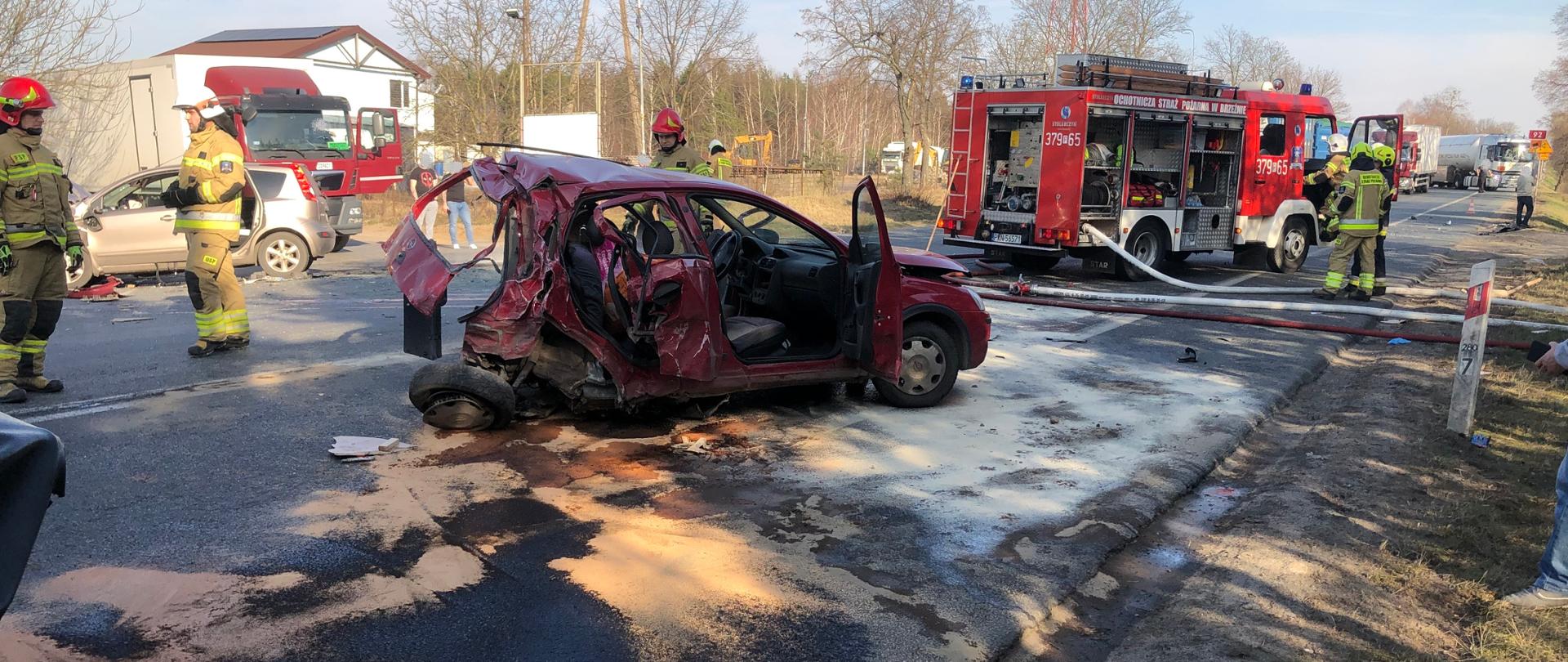 Na środku jezdni stoi uszkodzony samochód osobowy marki Opel. Drugi samochód marki Nissan stoi po drugiej stronie. Samochód strażacki stoi na środku jezdni i ma rozwinięte linie wężowe. Strażacy stoją przy pojazdach. Na jezdni rozlane płyny eksploatacyjne. 
