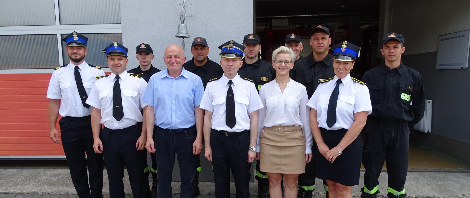 Zdjęcie wykonano w ciągu dnia na placu Komendy Powiatowej Państwowej Straży Pożarnej w Sępólnie Krajeńskim. Strażacy Państwowej Straży Pożarnej ubrani w mundury koszarowe, Komendant Powiatowej Straży Pożarnej oraz pracownicy biurowi ubrani w mundury wyjściowe pozują do zdjęcia przed budynkiem Komendy Powiatowej Straży Pożarnej.