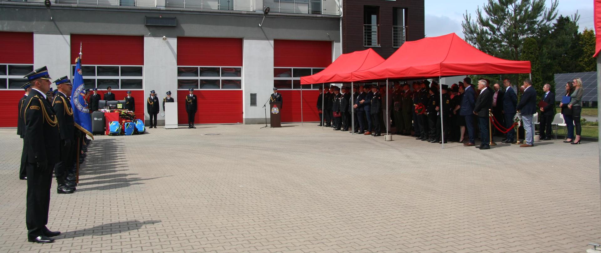 Na tle budynku po lewej stronie dowódca pododdziału oraz Sztandar Komendy Powiatowej Państwowej Straży Pożarnej w Lublińcu oraz pododdział strażaków w środkowej części pod baldachimem grupa zaproszonych gości.