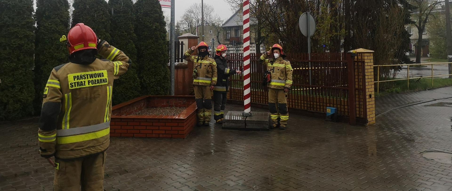 Na drugim planie widoczni strażacy dokonujący uroczystego podniesienia flagi Rzeczypospolitej Polskiej. Na pierwszym planie, po lewej stronie strażak salutujący, zwrócony frontem do masztu.