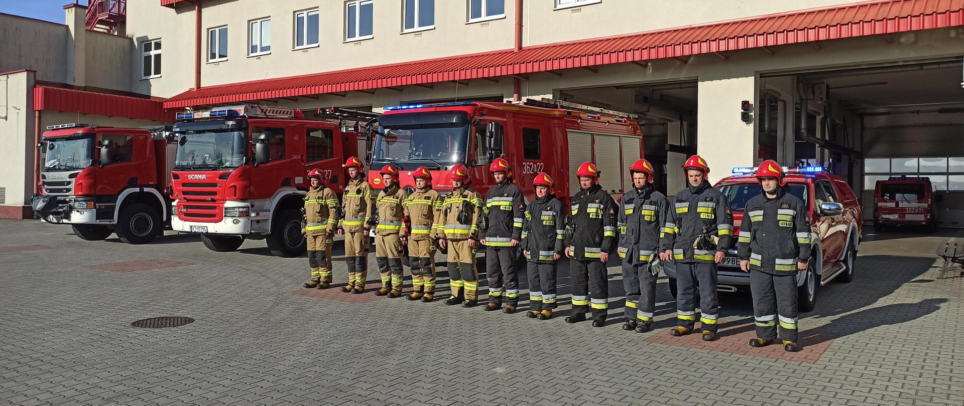 Na zdjęciu widoczni są strażacy Jednostki Ratowniczo-Gaśniczej nr 2 w Tarnowie stojący w czasie apelu upamiętniającego strażaków ukraińskich. Strażacy umundurowani są w ubrania bojowe, stoją na tle garażu, z tyłu widoczne samochody bojowe.