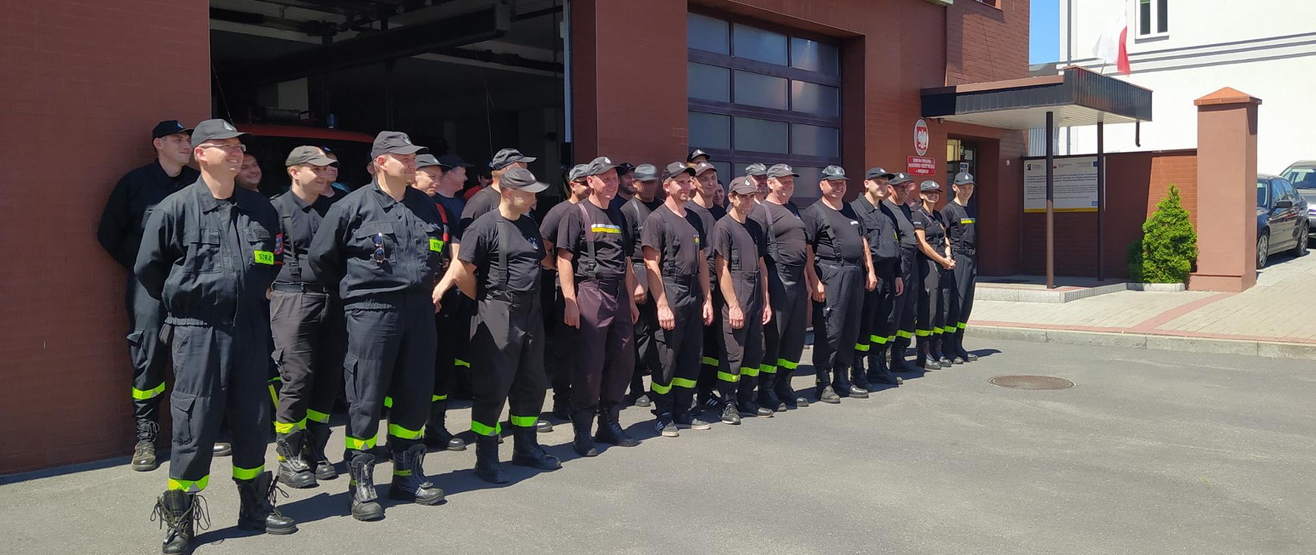  na tle budynku w trzech rzędach stoją druhowie OSP w czarnych ubraniach strażackich. Zdjęcie wykonane na zewnątrz w słoneczną pogodę