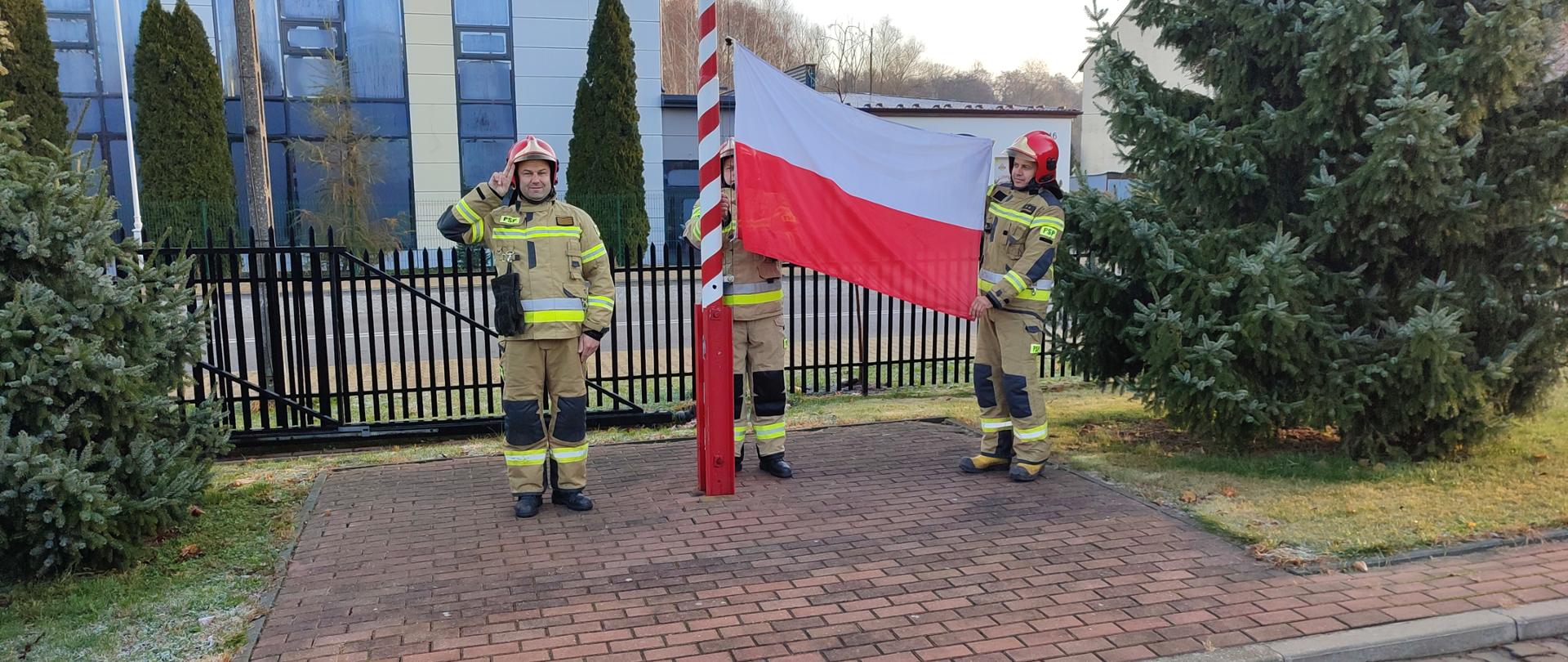Strażacy ubrani w ubrania specjalne podczas zmiany służbowej dokonują uroczystego podniesienia flagi państwowej na placu Komendy.