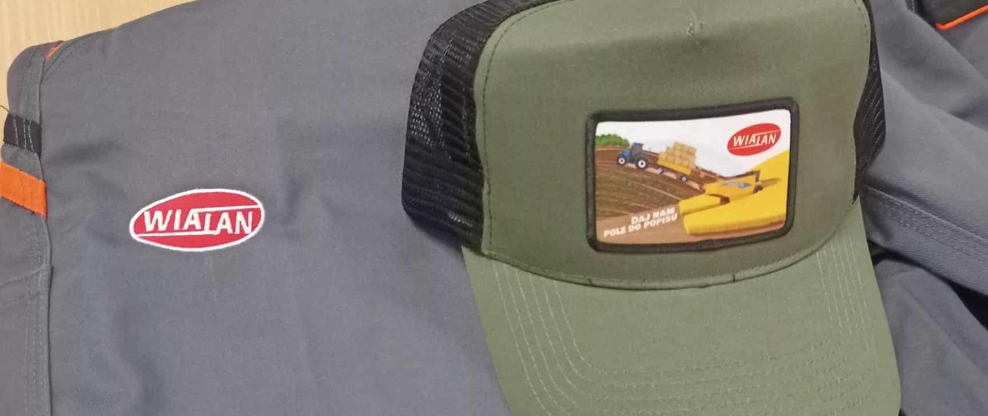 Zbliżenie na szarą odzież roboczą oraz zieloną czapkę z daszkiem typu „trucker”. Na obu elementach widoczne jest czerwono-białe logo firmy „Wialan”.