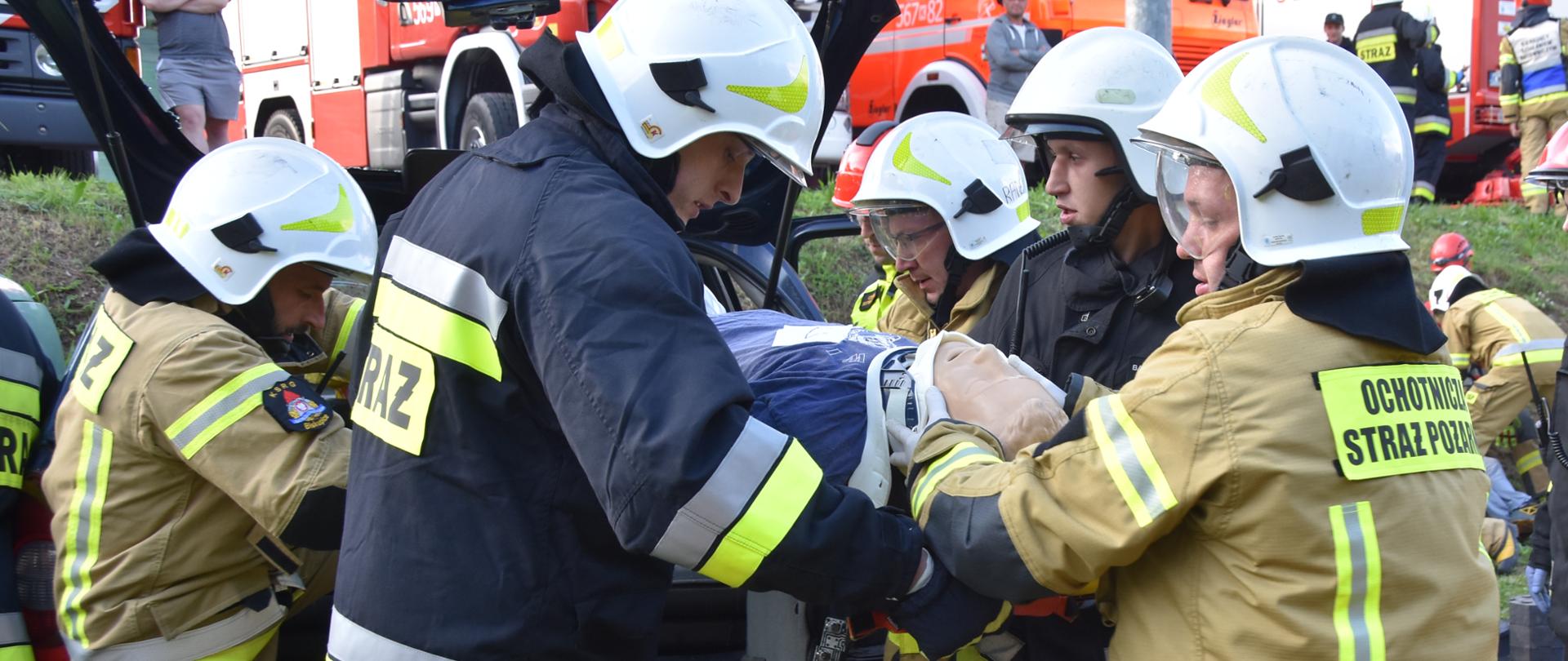 Na zdjęciu strażacy w ubraniach specjalnych, którzy ewakuują poszkodowanego (manekina) z wraku samochodu osobowego.