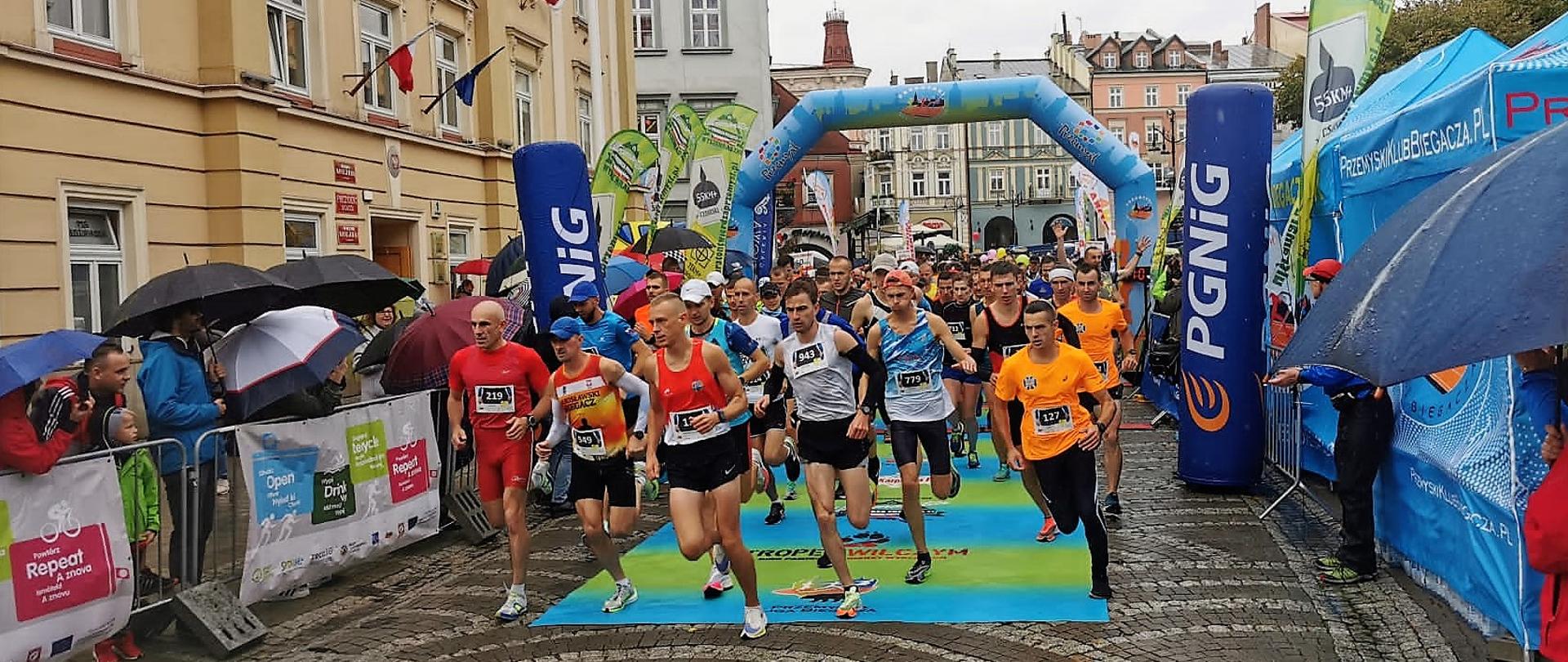 Fragment wyścigu. Uczestnicy biegnący w grupie po bruku na tle linii startu
