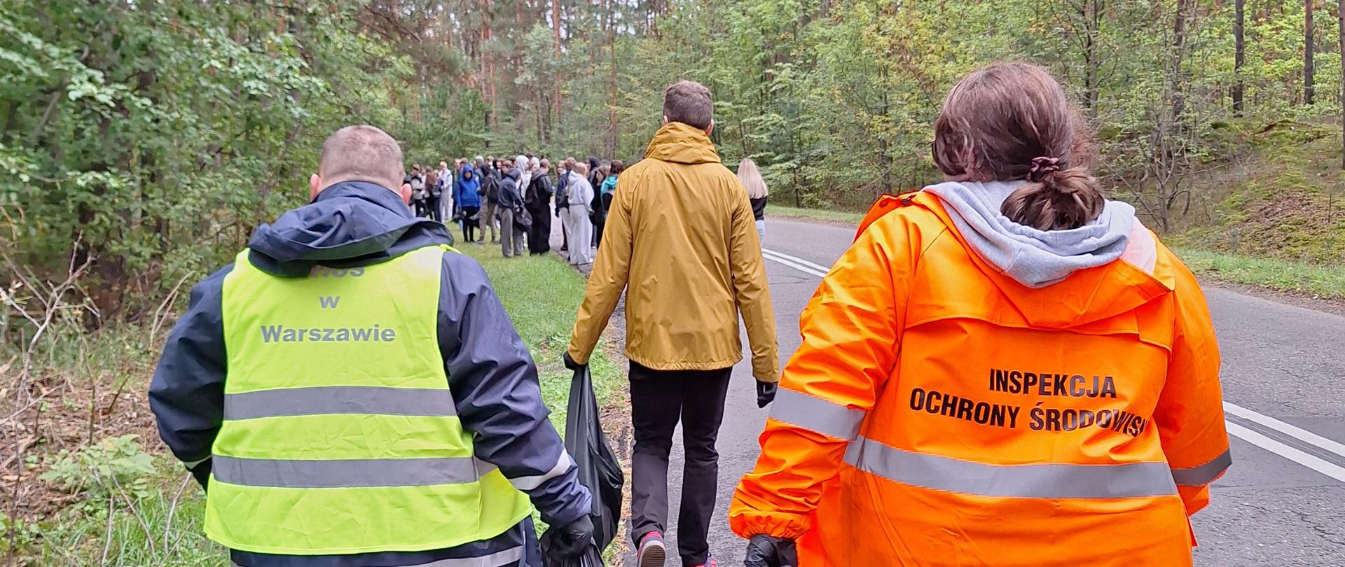 Grupa ludzi, w tym dwoje inspektorów WIOŚ w Warszawie idzie asfaltową drogą przez las. Jeden z inspektorów niesie w dłoni worek na śmieci.