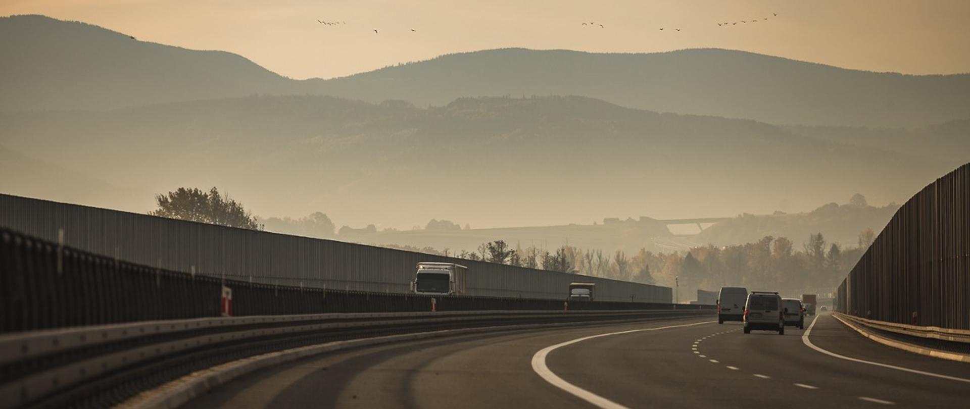 Fotografia dwujezdniowej drogi ekspresowa S1. W tle widoczne góry. W górnej części fotografii widoczne niebo z kluczem przelatujących ptaków. Po jezdniach w obu kierunkach poruszają se pojazdy. 