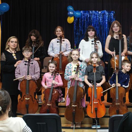 Zdjęcie przedstawia grupę uczniów stojących na scenie z wiolonczelami. W tle karnawałowe dekoracje.