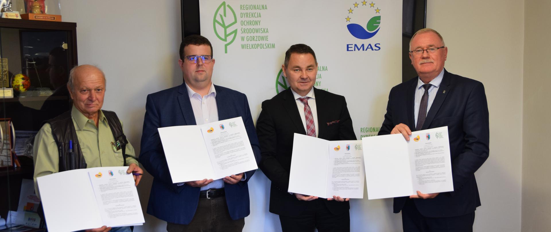 Czterech mężczyzn pozuje do zdjęcia. W rękach trzymają teczki, a w nich znajdują się zapisane kartki papieru.