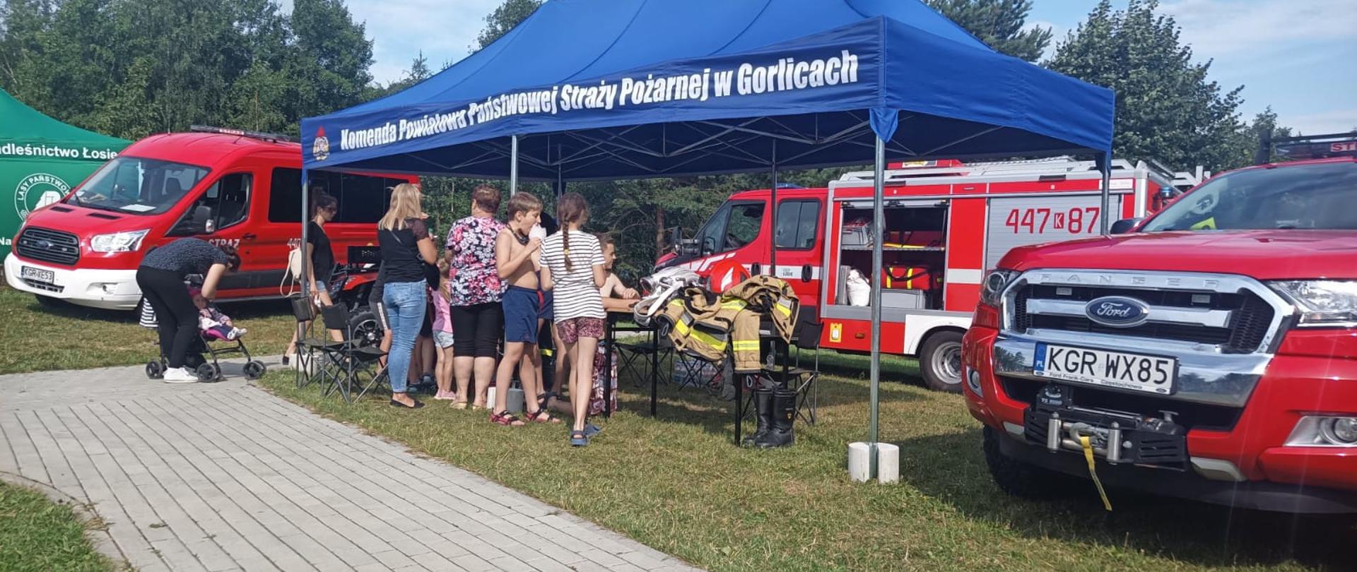Na zdjęciu widoczna grupa dzieci i osób dorosłych uczestniczących w pikniku rodzinnym nad zalewem Klimkówka przed stanowiskiem wystawienniczym Komendy Powiatowej Państwowej Straży Pożarnej w Gorlicach. W którym strażacy prowadzili prelekcje i konkursy z zakresu bezpieczeństwa przeciwpożarowego i bezpieczeństwa nad wodą. W tle widoczne samochody pożarnicze koloru czerwonego.