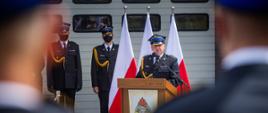 Na zdjęciu widoczny Komendant Powiatowy PSP w Gorlicach w umundurowaniu galowym granatowym w czapce rogatywce przy mównicy podczas przemówienia z okazji Powiatowych obchodów Dnia Strażaka. W tle trzy flagi biało czerwone i strażacy w umundurowaniu galowym. 