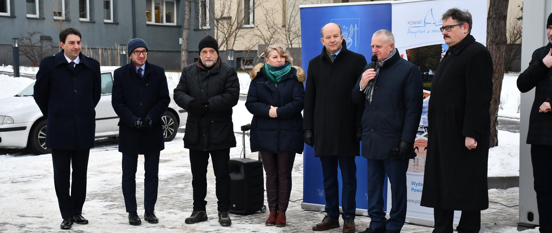 Na zdjęciu znajduje się osiem osób - kobieta i siedmiu mężczyzn. Osoby te stoją w rzędzie. Jeden z mężczyzn ma w ręku mikrofon, będąc w trakcie wystąpienia publicznego. Zdjęcie wykonane zostało na dworze, w zimowej scenerii. 
