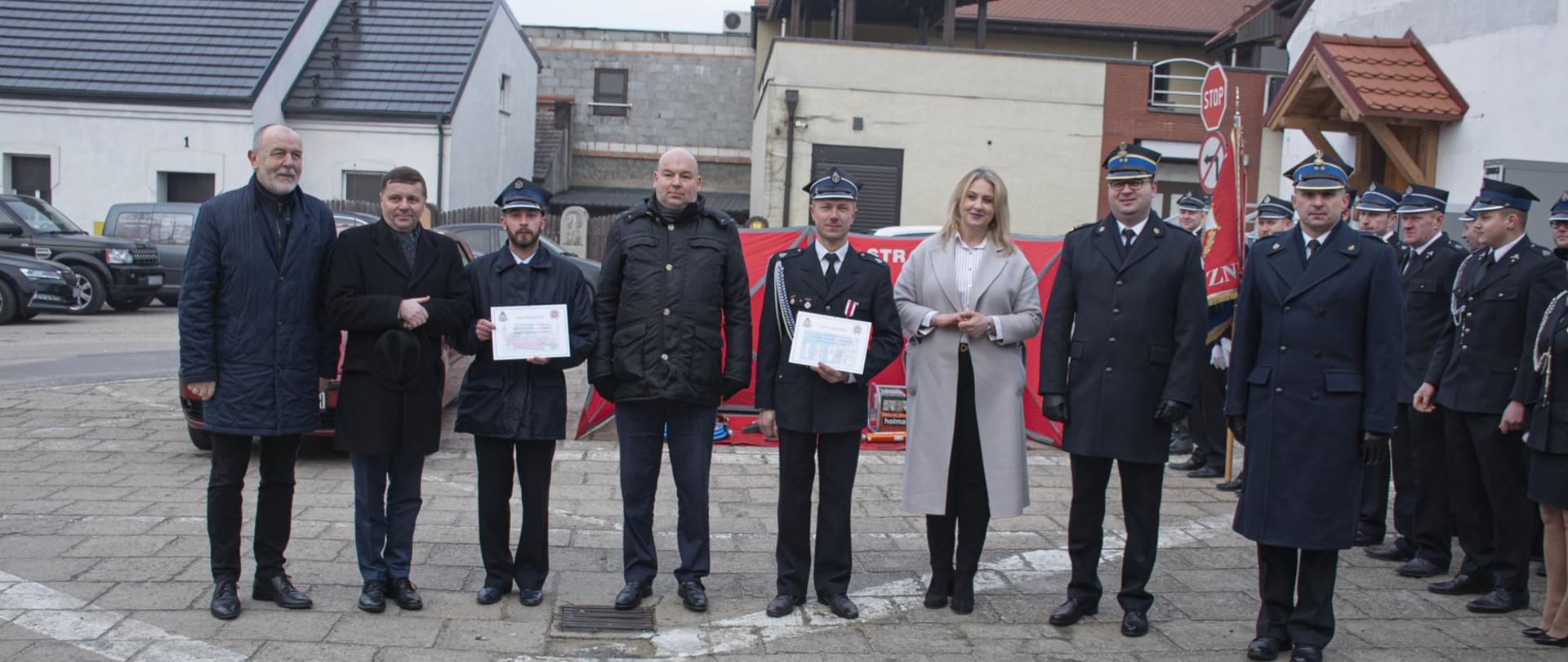 Zdjęcie przedstawia osoby, które przekazywały sprzęt jednostką OSP podczas wspólnego zdjęcia na tle przekazywanego sprzętu.