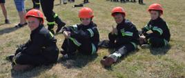 Zawody gminne w Śląskowie. Na placu ćwiczeń siedzi czterech członków Młodzieżowej Drużyny Pożarniczej. Na głowach maja czerwone hełmy. W tle inni zawodnicy oraz kibice.