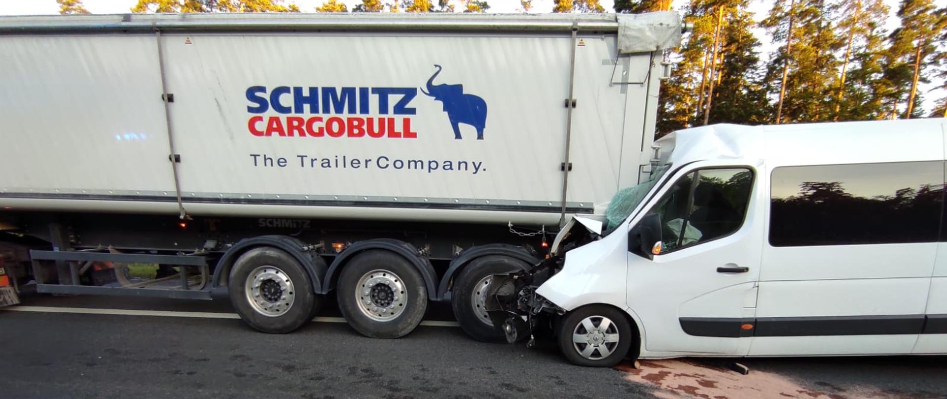 Zdjęcie pokazuje busa osobowego wbitego w tył naczepy przewożącej kruszywo. Siła uderzenia była tak duża, że tył naczepy przebił przednią szybę i znajduje się tuż przed twarzą kierowcy i pasażera busa. Z busa wylały się płyny eksploatacyjne stąd tuż przed nim widać różowy sorbent, którym strażacy posypali jezdnię celem sorpcji i neutralizacji substancji. W tle widać drzewa.