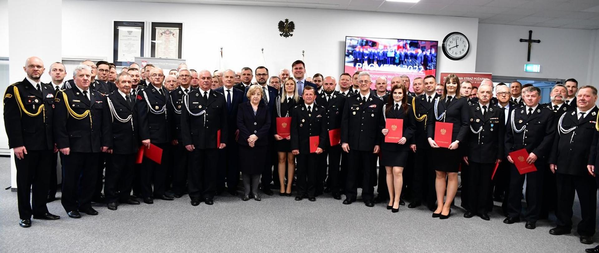 Zdjęcie przedstawia uczestników uroczystości ustawionych w półksiężyc. W tle ekran monitora, zegar i godło.