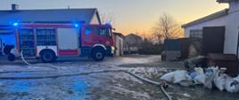 Na zdjęciu widać wóz strażacki zaparkowany między budynkami gospodarczymi. Ziemia jest miejscami oblodzona i pokryta resztkami śniegu. Na samochodzie strażackim świecą się niebieskie światła sygnalizacyjne, a z boku wozu widać otwarte skrytki ze sprzętem gaśniczym. Na ziemi widać rozłożone odcinki węży strażackich, jeden z nich podłączony do rozdzielacza. Obok znajdują się worki oraz różne przedmioty gospodarcze. W tle widać budynki, ogrodzenie oraz drzewa.
