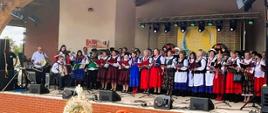 Zdjęcie przedstawia scenę plenerową, na której odbywa się występ artystyczny. Scena znajduje się pod dużym, drewnianym zadaszeniem. Na scenie występuje duża grupa osób ubranych w tradycyjne ludowe stroje, charakterystyczne dla regionu. Występ ma charakter chóralno-instrumentalny. 