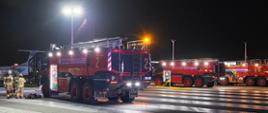 Kilka lotniskowych pojazdów ratowniczo-gaśniczych stoi w gotowości na drodze startowej podczas nocnych ćwiczeń służb ratowniczych. W tle widoczny jest samolot, a przy jednym z wozów strażacy przygotowują sprzęt wykorzystywany podczas symulacji zdarzenia lotniczego