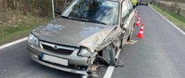 Zdjęcie przedstawia samochód osobowy stojący na pasie jezdni blokujący jeden pas ruchu. Samochód z uszkodzonym lewym przodem. Na zdjęciu widać również pachołki. 