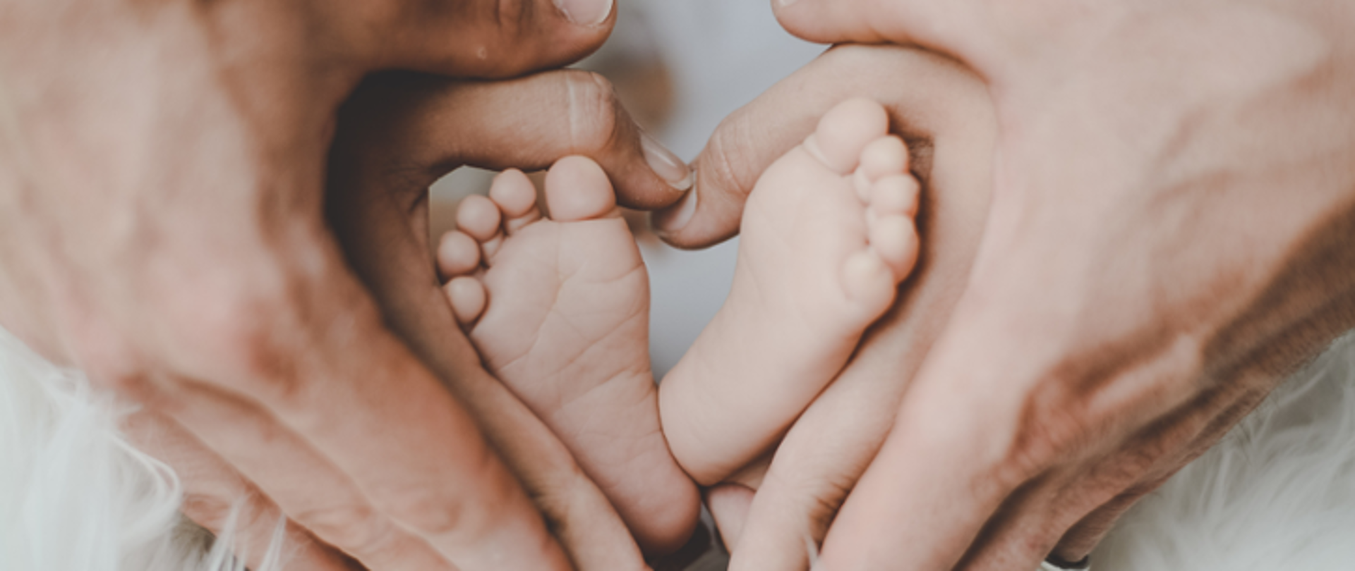 Na zdjęciu znajdują się stópki noworodka w objęciach dłoni kobiety i mężczyzny. 