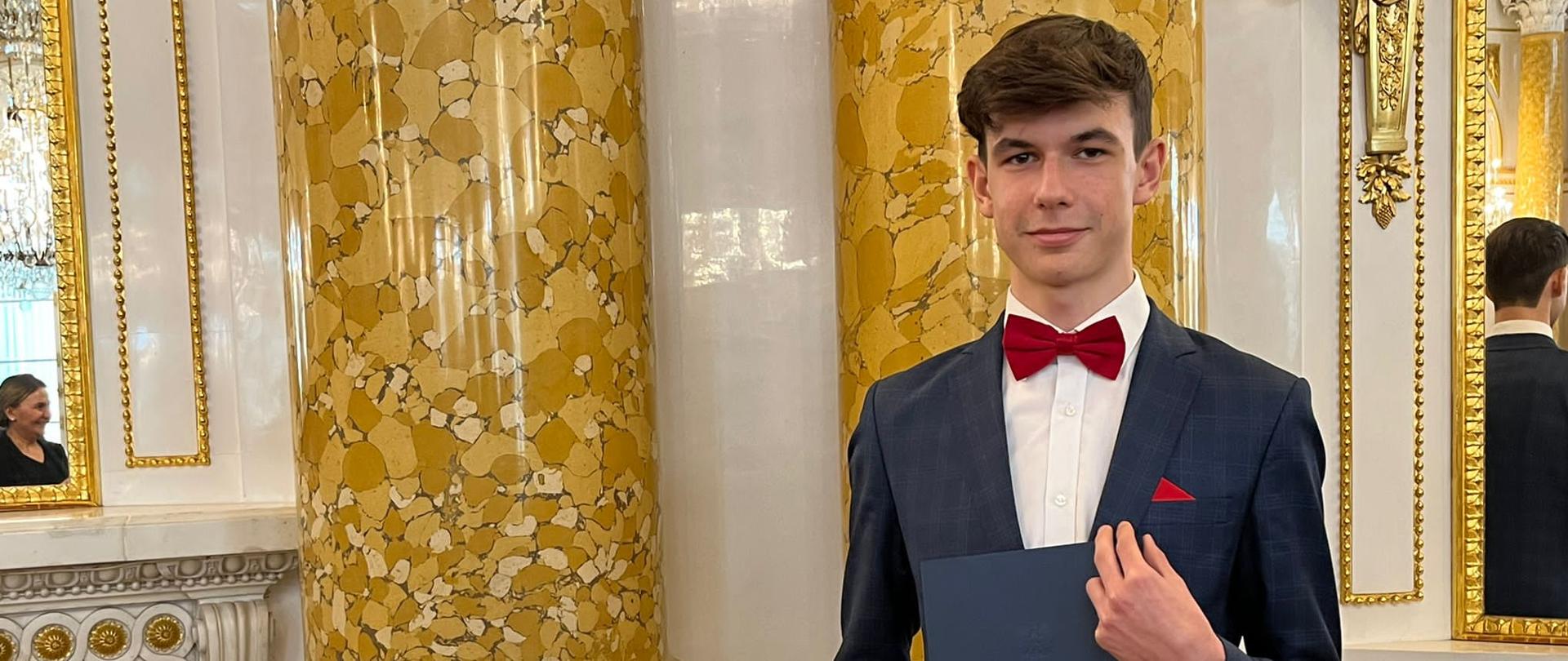 Bartosz Wódczak stoi w garniturze i trzyma teczkę ze stypendium, w tle kolumny.