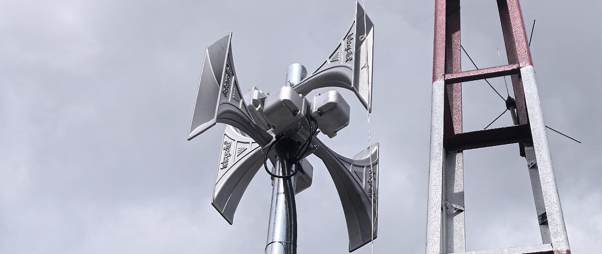 Zbliżenie na nowoczesną syrenę elektroniczną z kilkoma tubami zamontowaną na metalowym maszcie, obok wieży starego typu. W tle niebo z zachmurzeniem.