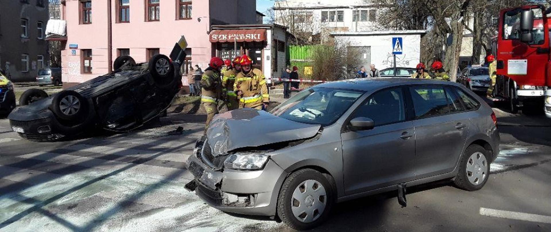 Dwa samochody osobowe po kolizji, jeden leżący na dachu, w tle strażacy oraz budynki wielorodzinny.