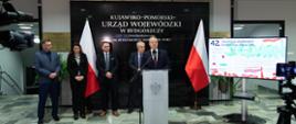 Jeden mężczyzna stojący przy mównicy i wypowiadający się. Za nim kobieta i trzech mężczyzn