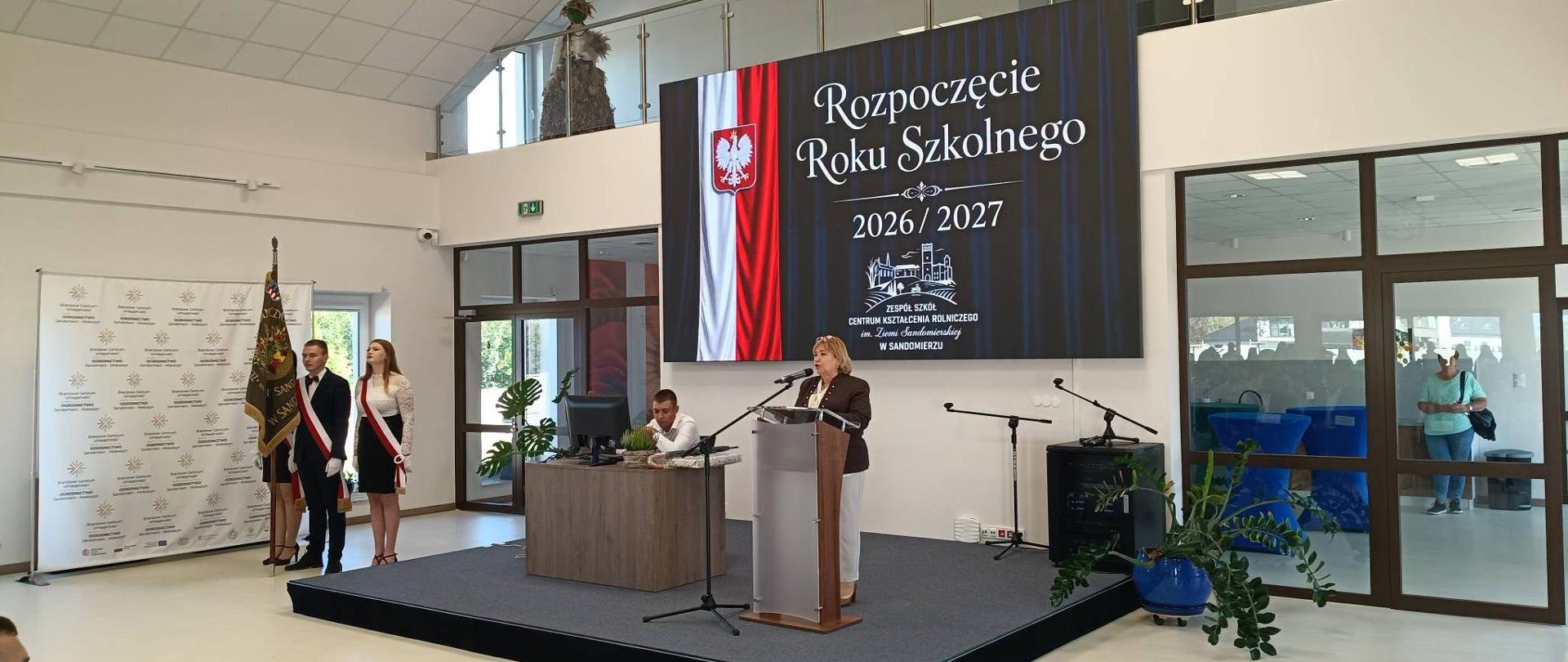 Dyrektor szkoły stojąca przy mównicy na niewielkim podwyższeniu. Z tyłu ekran z napisem Rozpoczęcie roku szkolnego 2026/2027. Po lewej dwie uczennice i jeden uczeń ze sztandarem szkoły. Wszystkie osoby w strojach galowych.