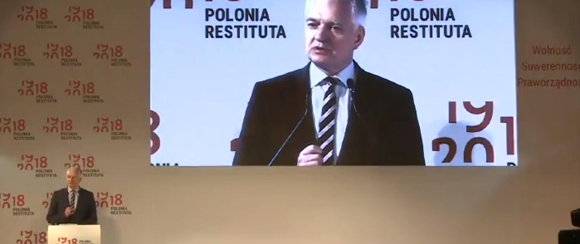 Minister Gowin stoi za mównicą, za nim ściana na której wyświetlany jest jego obraz