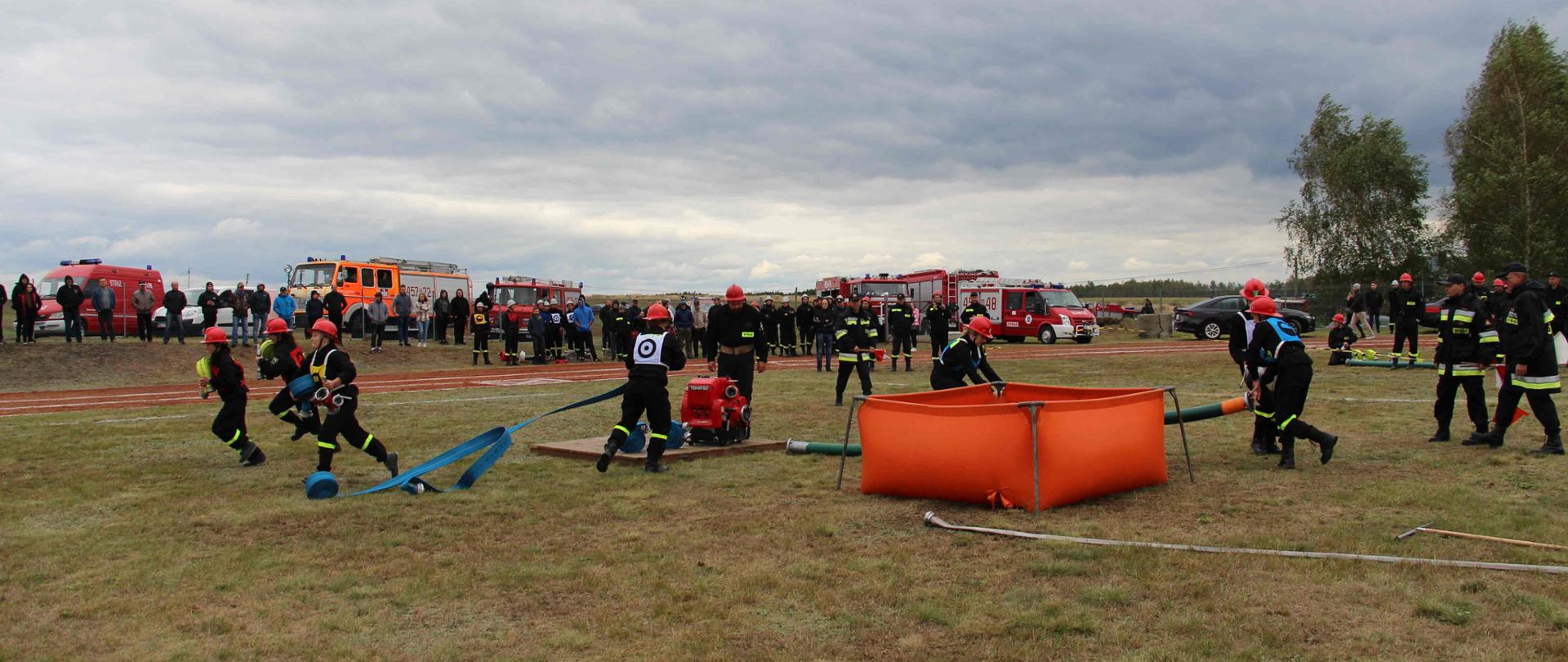 Na stadionie sportowym na trawie rozgrywane są zawody sportowo-pożarnicze – ćwiczenie bojowe. Na podeście stoi motopompa, w niewielkiej odległości od niej pomarańczowy zbiornik z wodą. Przy nich kilka druhen w czarnych ubraniach koszarowych rozkładają sprzęt. W tle osoby obserwujące zawody oraz samochody strażackie.