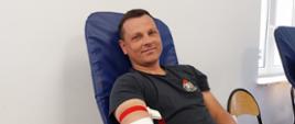 Na zajęciu widoczny strażak w czarnej koszulce i w czarnych spodniach siedzący na fotelu na sali oddający krew podczas akcji zbiórki krwi zorganizowanej przez Klub Honorowych Dawców Krwi Polskiego Czerwonego Krzyża przy Komendzie Powiatowej Państwowej Straży Pożarnej w Gorlicach