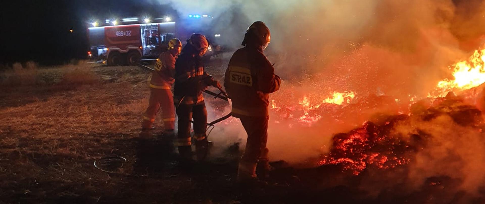 Po prawej stronie płonący stóg słomy. Po środku pracujący strażacy. W tle samochód pożarniczy 