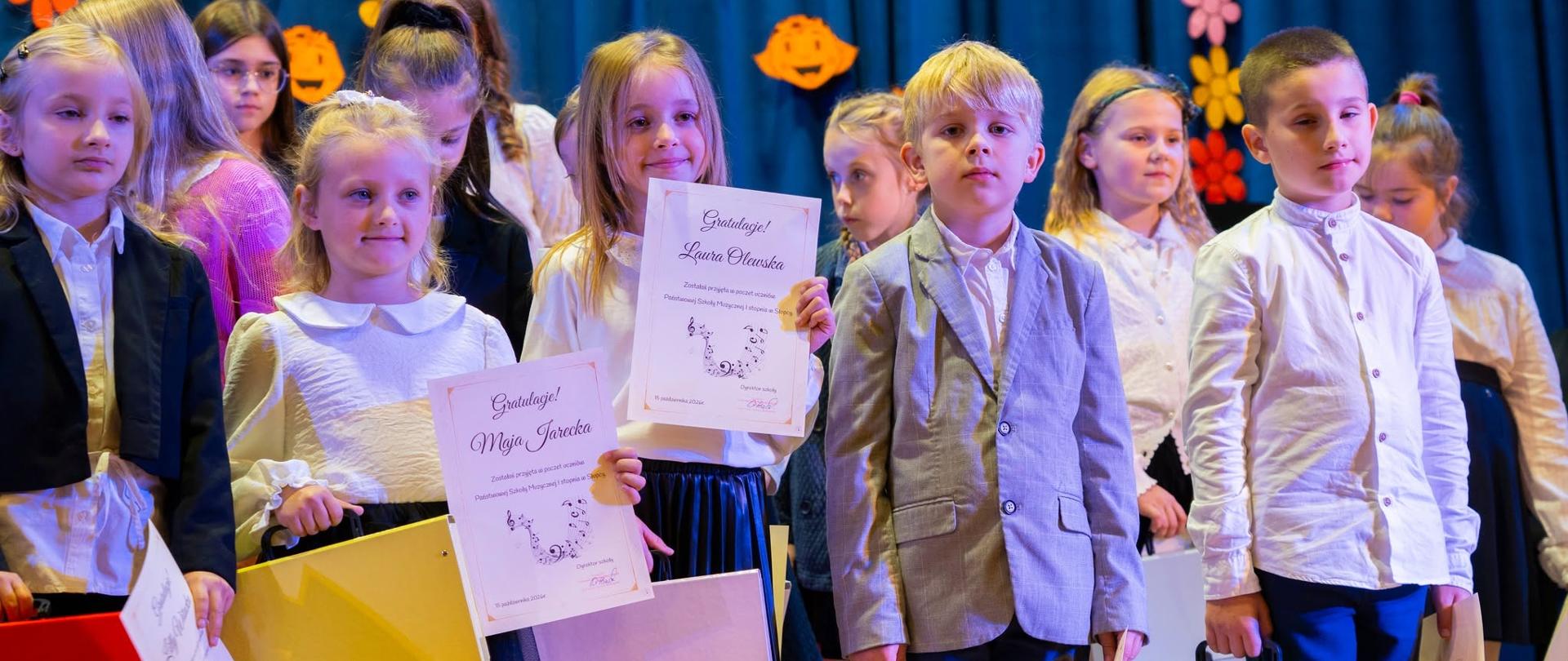 Grupa elegancko ubranych dzieci stojących na scenie podczas uroczystości szkolnej, trzymających dyplomy gratulacyjne i kolorowe teczki.